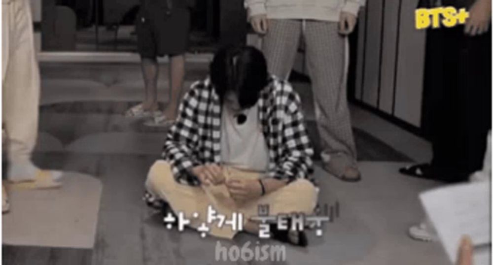 a man in a plaid shirt sits on the floor in front of a sign that says bts+