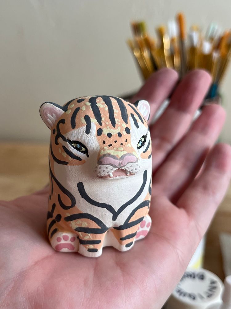 An unglazed handmade ceramic tiger sitting in the palm of a hand.