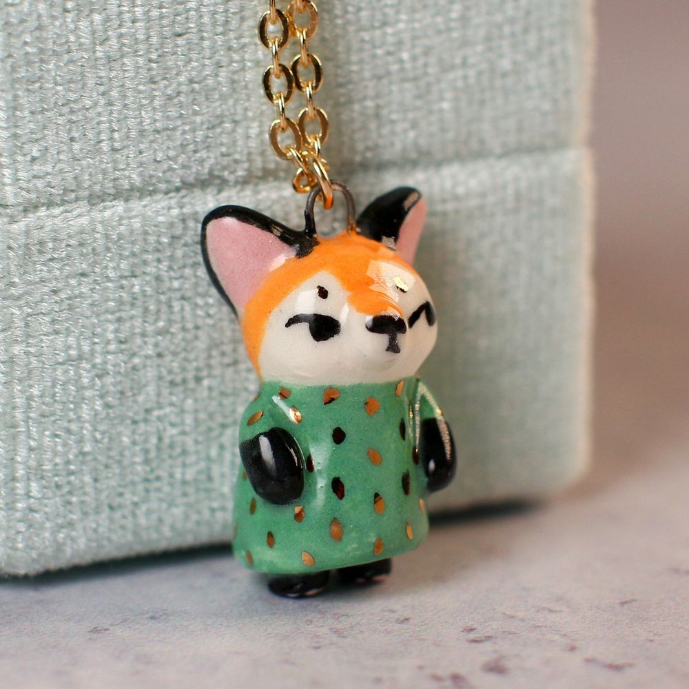 Handmade ceramic fox pendant wearing a green dress with gold polka-dots glances sideways to something out of frame. 