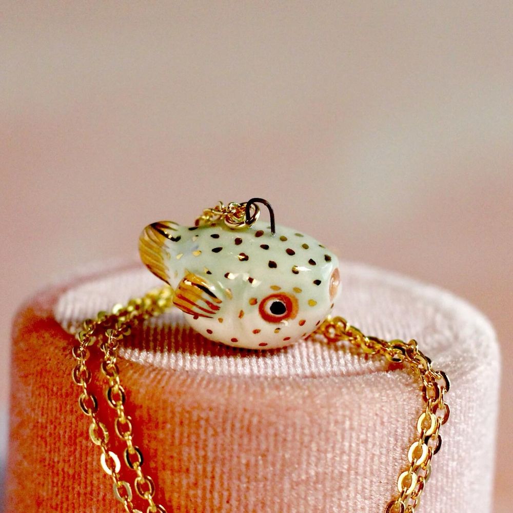 A pale mint green ceramic puffer fish pendant with mustard fins and gold details sits on a pink velvet ring box.