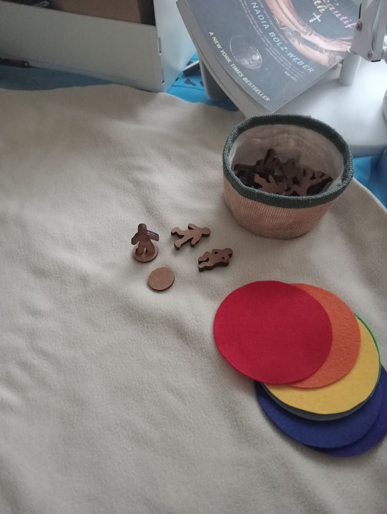 A small Hessian basket containing wooden figures with a few wooden figures in different poses and some rainbow coloured felt circles on a large cream fabric circle