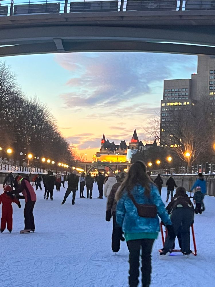 ottawa frozen canal with people ice skating and lights and parliament building in the distance, with a pretty sunset