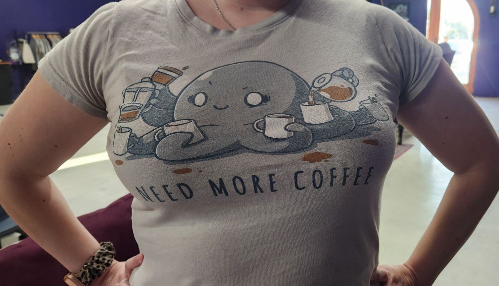 Someone wearing a Grey tee featuring a friendly octopus. Each tentacle is holding either a cup of coffee or pouring coffee from a carafe into a mug. The caption reads "Need more coffee."  Me too, sweet Octopus. Me too.