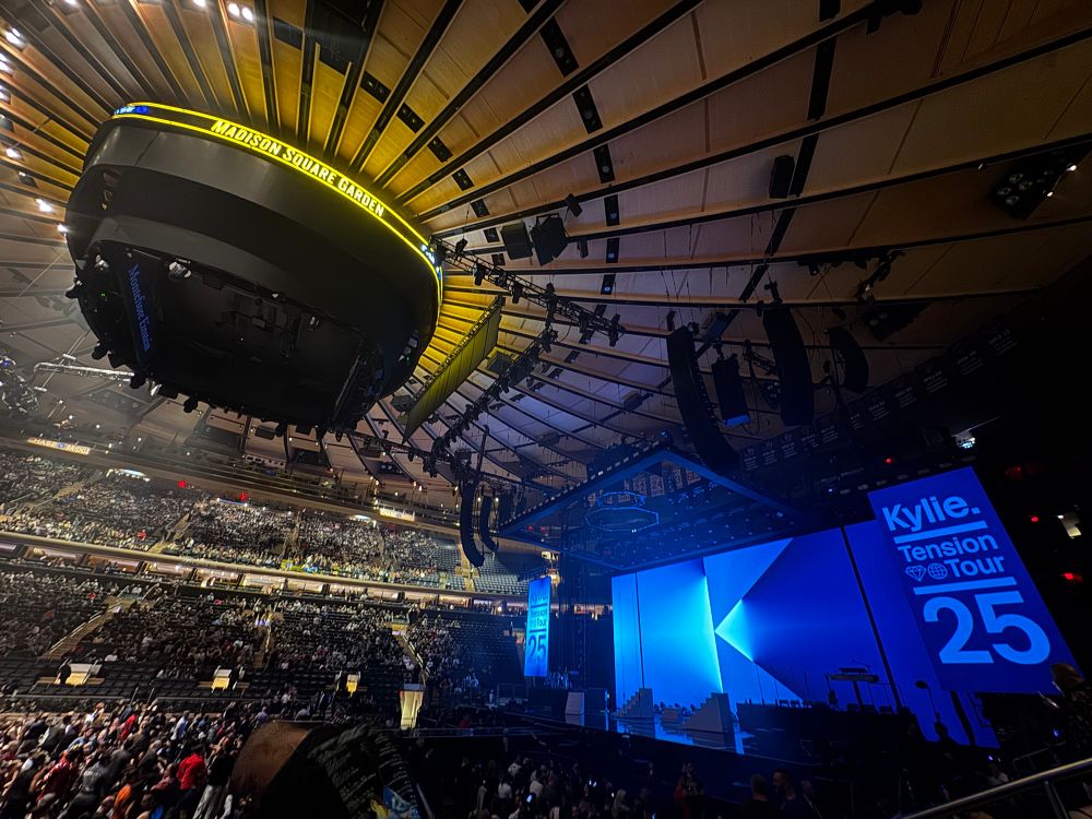 Picture of the stage and ceiling, you can see Jumbotron that says “Madison Square Garden”.  a crowd is filling in.  the stage has a large center screen with various blue triangles shaped to make a ‘K’.  on the right of that, a smaller screen that says “Kylie. Tension Tour 25”