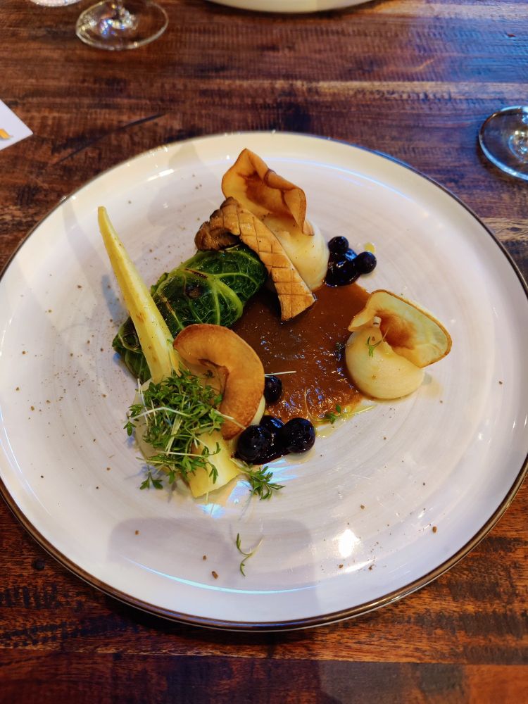 Zu sehen ist ein Teller mit Pastinakenpüree, Blaubeeren und einer Wirsingroulade gefüllt mit Steinpilzen und Kräuterseitlingen. Dazu ein wenig Kresse und Pastinaken-Chips.