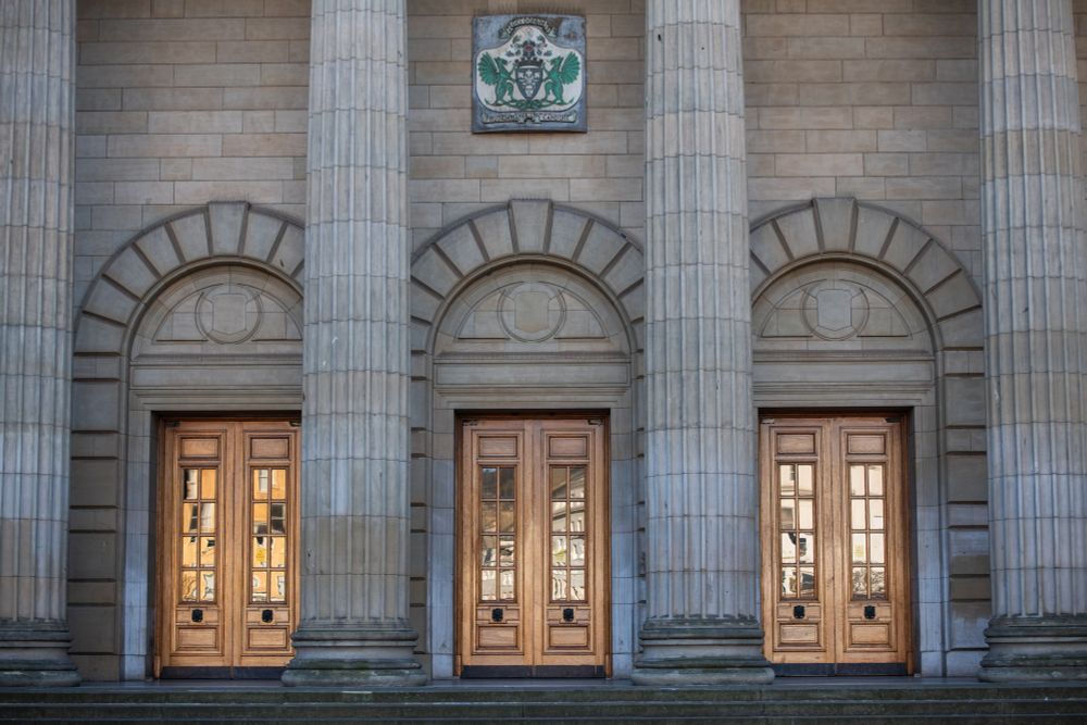 3 grand doorways with arches above are flanked by tall doric columns. Above the doors is a city crest depicting a large urn of lily's. 