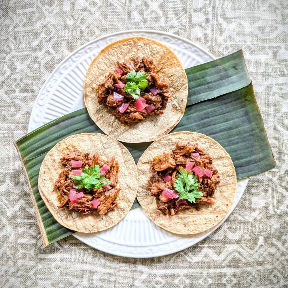 Three tacos on a plate with a banana leaf, filled with meat, topped with pickled onions and cilantro.