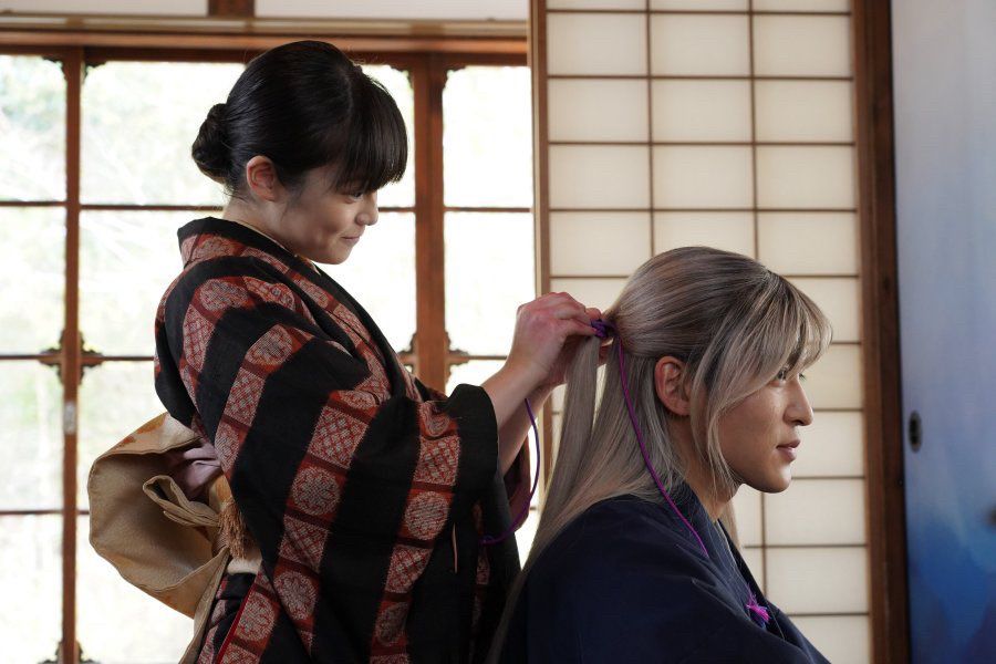 A still from the film "As Long As We Both Shall Live" (2023), a live action adaptation of the manga & light novel “My Happy Marriage”. Miyo in a kimono smiles softly as she ties her fiancé Kiyoka’s long silver hair.