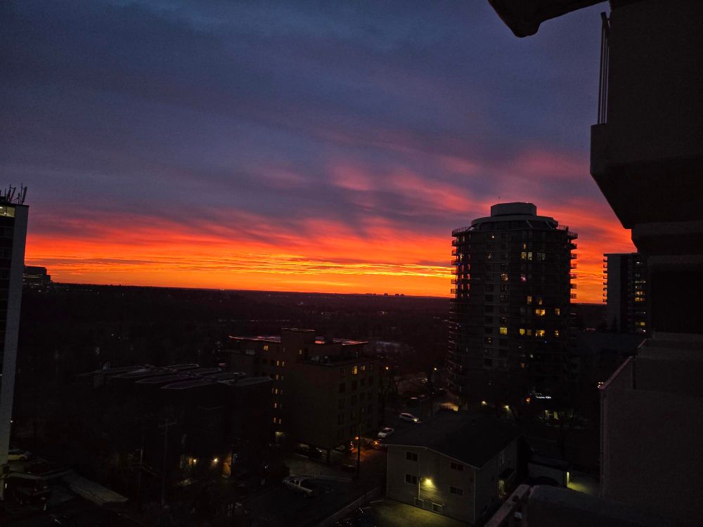 bright orange stripe with yellow graduations under a purple clouded sky with sone pink swooshes (over the Edmonton River Valley. a dark building with a few lights on stands in front