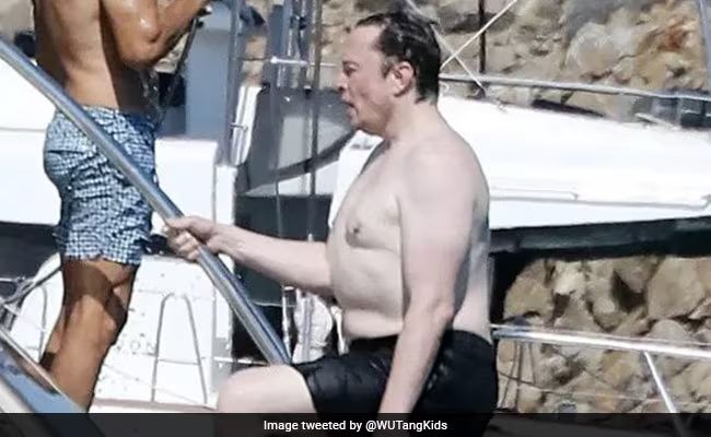 Ein Foto von Elon Musk, wie er in einer schwarzen Badehode die Leiter einer Jacht hinaufsteigt.