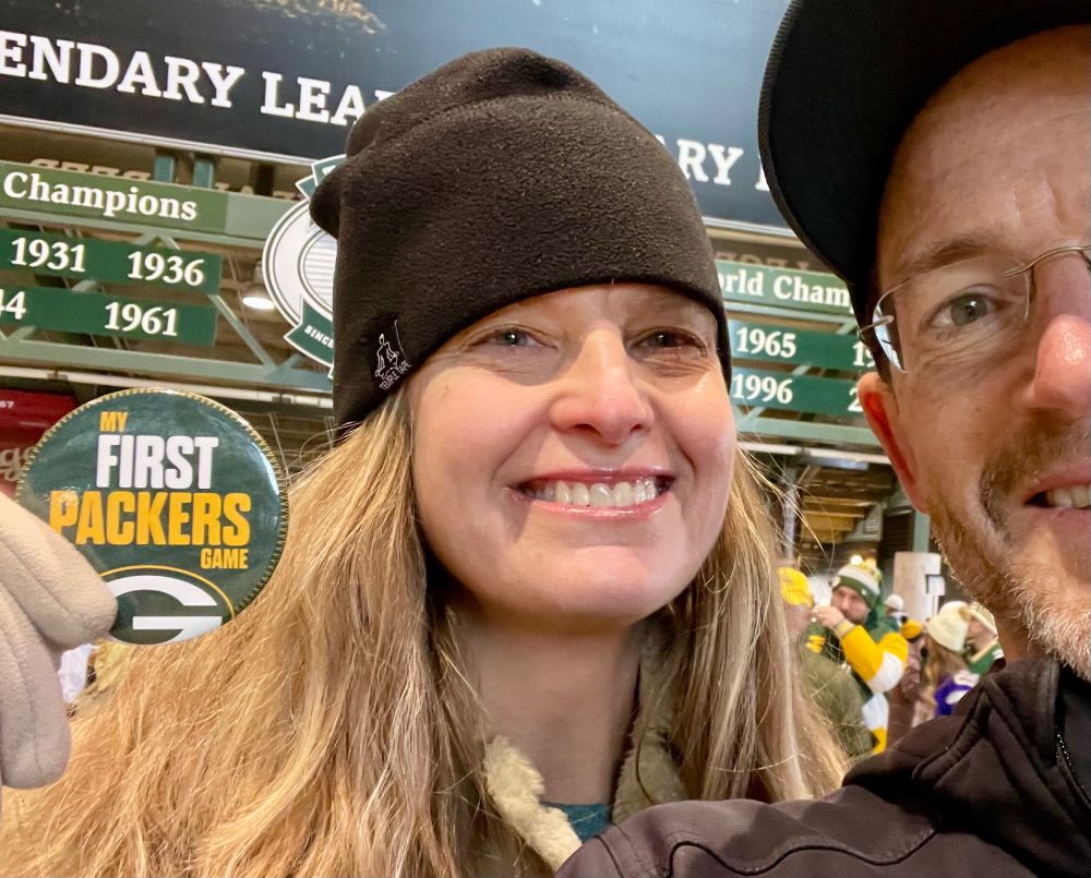 Blonde-haired woman with a black cloth cap holding up a button that says "My First Packers Game"