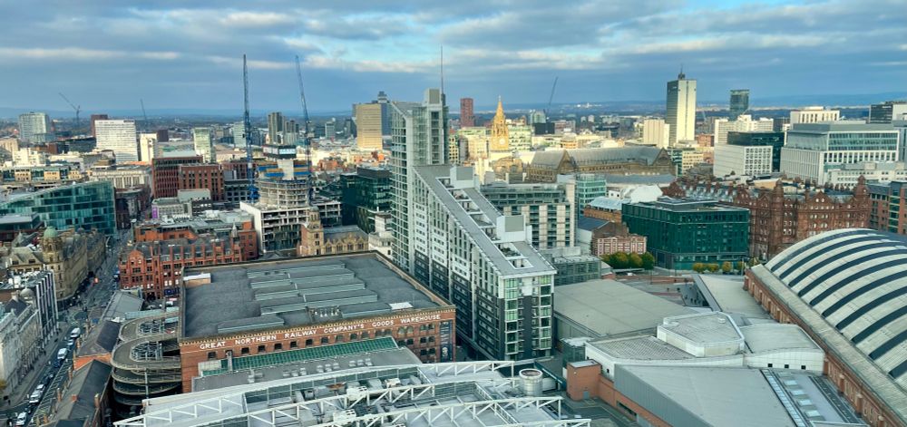 View of central Manchester UK under a partly cloudy sky from a 23rd floor, offering a broad view of many buildings into the distance. A line of sunshine illuminates a line of buildings near the horizon.