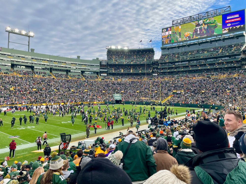 Fans applauding the end of a Packers win on Lambeau Field under a cloudy sky. The teams are meeting together in the middle of the field.