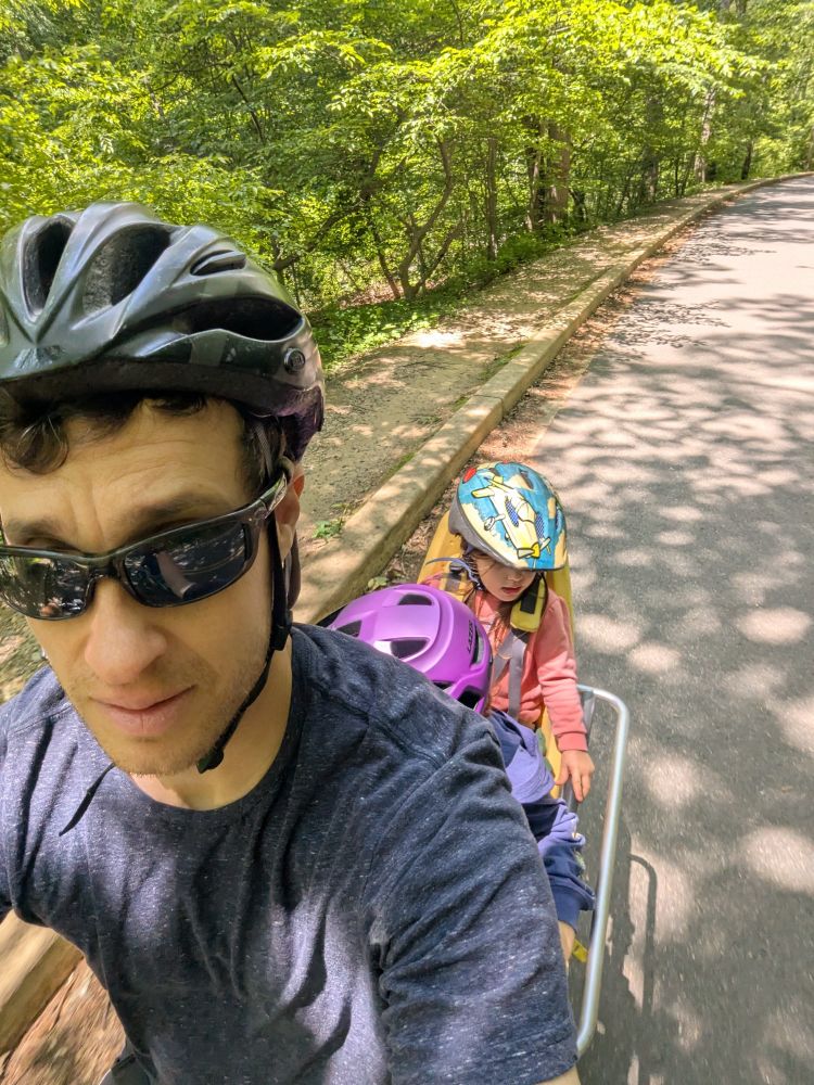 A man on a cargo bike with two young children, riding on a road through Rock Creek Park surrounded by lush trees.