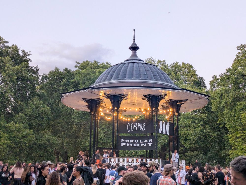 Performance by Show me the body at the Southwark Park Bandstand. With banners saying Popular Front, Corpus and  a banner saying "Free OLA" wrapped around it.