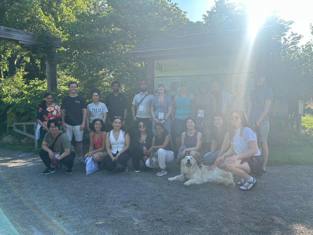 A group of smiling attendees posing in front of a trailhead sign with our new friend the golden retriever! The sun is also photobombing us. 