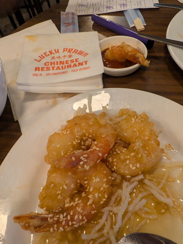A close-up, high-angle shot shows three fried prawns coated in a honey glaze and sesame seeds, served over fried white noodles on a white plate. A spoon rests in the lower right corner. In the background, a white napkin with "LUCKY PRAWN 美味中餐 CHINESE RESTAURANT DINE IN • AIR CONDITIONED • FULLY LICENSED" printed in orange ink, a small bowl of dipping sauce with Vinh's half eaten prawn, a purple pen, and an 80s music bingo sheet (just because) are on a dark wooden table.
