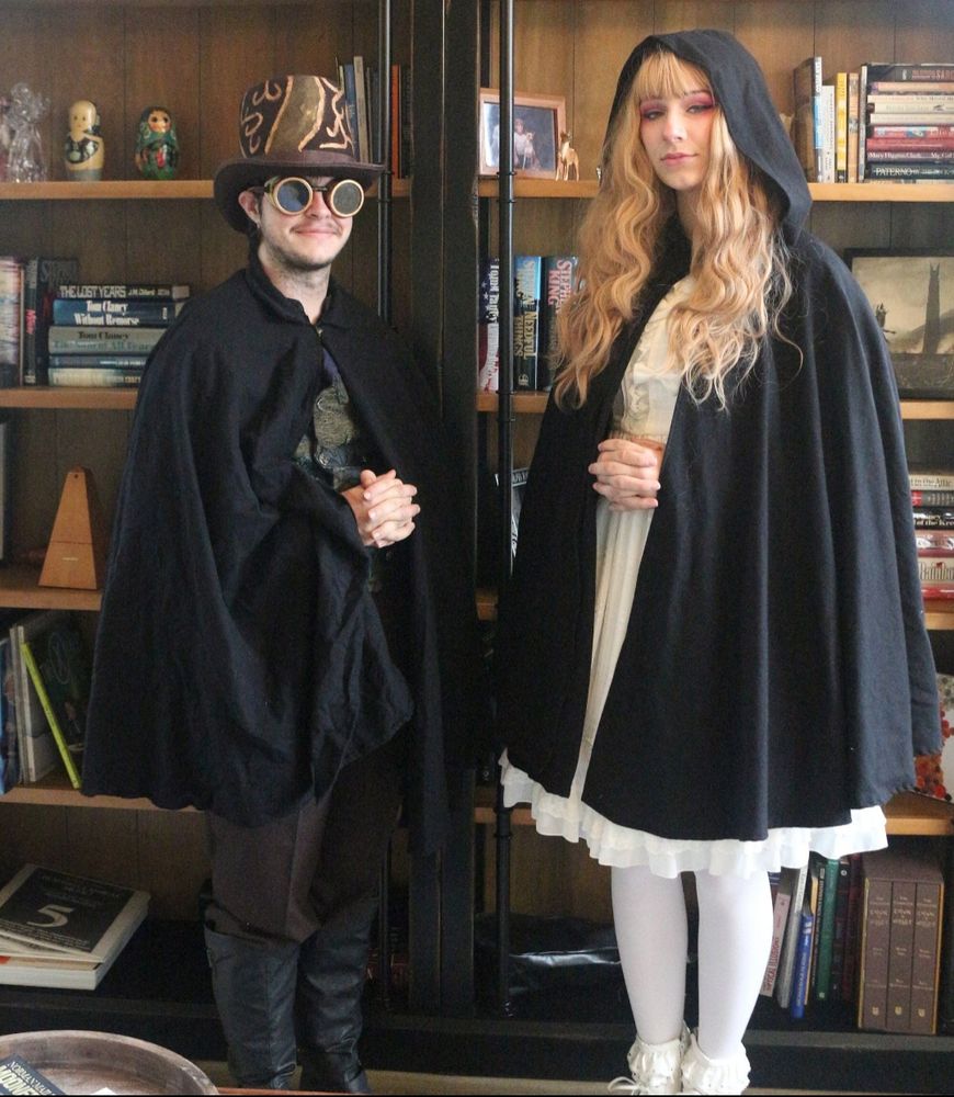 Two people wearing black capes in front of a bookshelf filled with books.