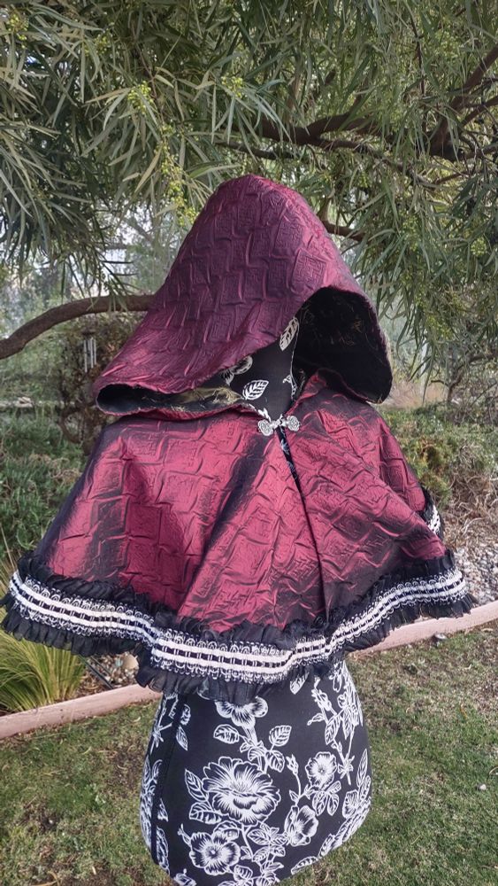 Red cape with textured, silk like fabric. Frilly, black and white trim sewn to the bottom of cape.
