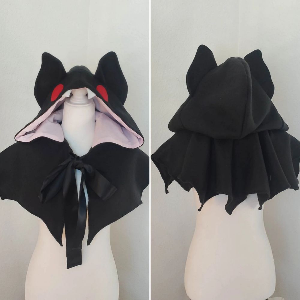 Fleece cape designed to look like a bat. Outer fabric is black fleece. The hood has big bat ears and red eyes. Inner lining is pink cotton. Black ribbon is used to close the cape.