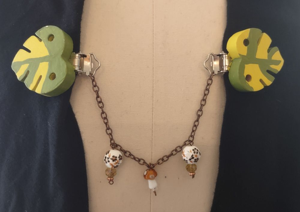 Cape clasp with monstera leaf base. Copper chain connects the two leafs. Ceramic pottery like beads and a brown, spotted glass mushroom hang from the chain.