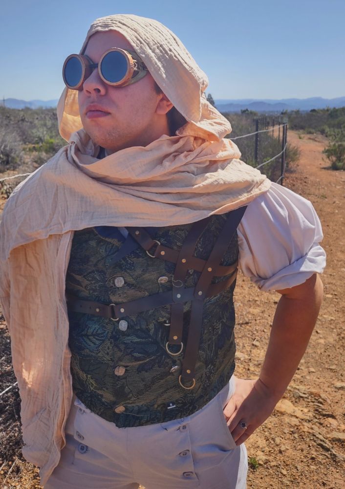 Man dressed in steampunk pants, vest, and shirt. He wears goggles and his head is wrapped in a lightweight gauze scarf to keep himself out of the sun.