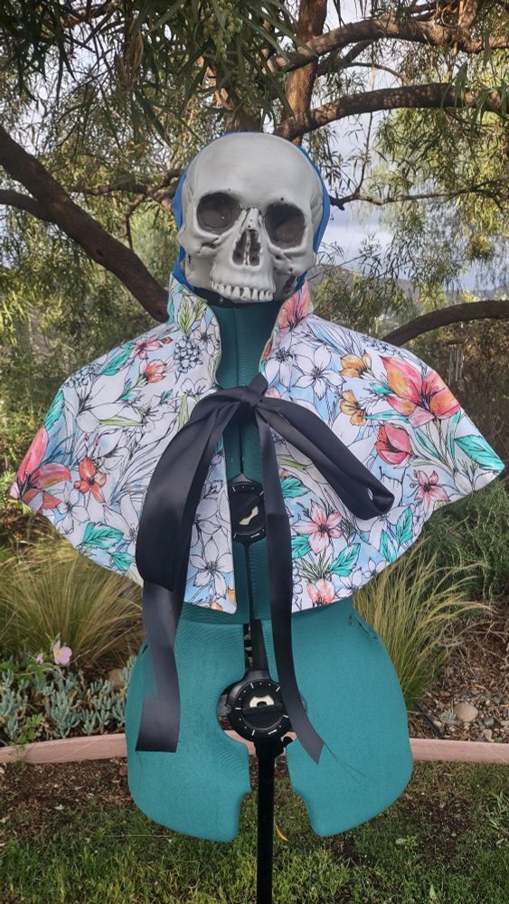 Dress form with a plastic skull taped to it wearing a short cape. Cape has a cute floral print on it. 