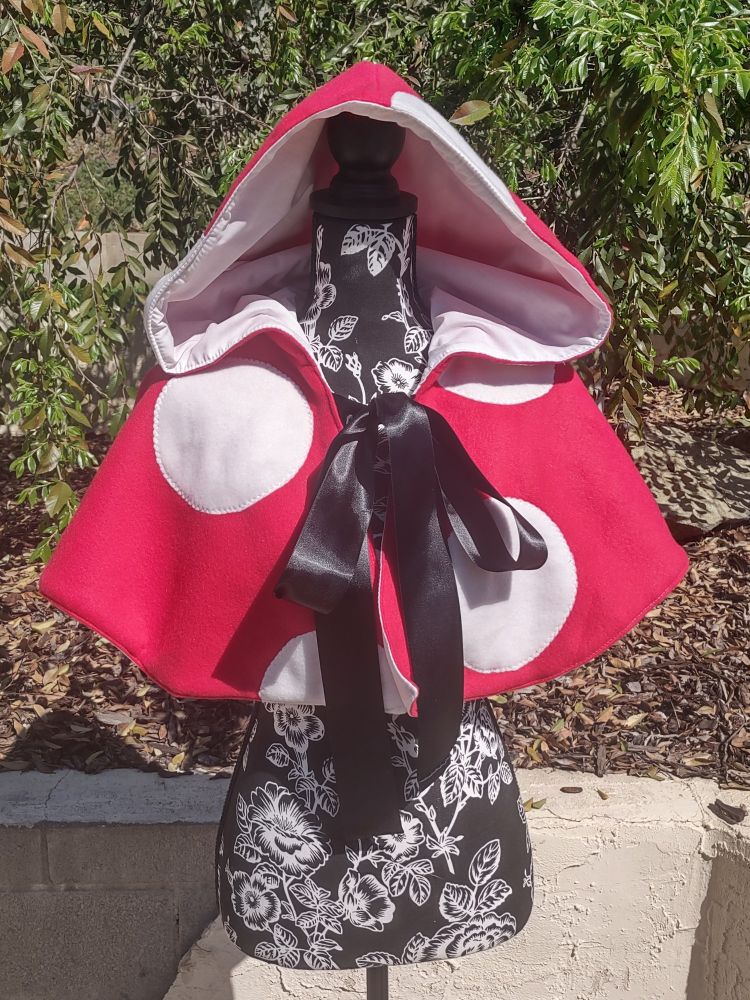 Mannequin with a mushroom cape on it. Mushroom cape goes to about the middle of the torso. Cape is red with white spots on it.