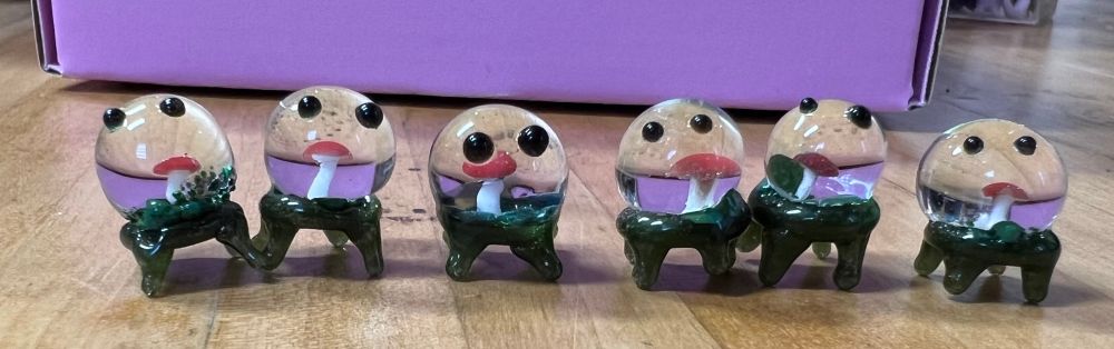 Six lil mushroom droplet guys made of borosilicate glass stand in a row on a wooden table. Each one looks like a water droplet encasing a tiny amanita mushroom with shiny black eyes (on the droplet) and mossy legs. They all vary a bit from eachother