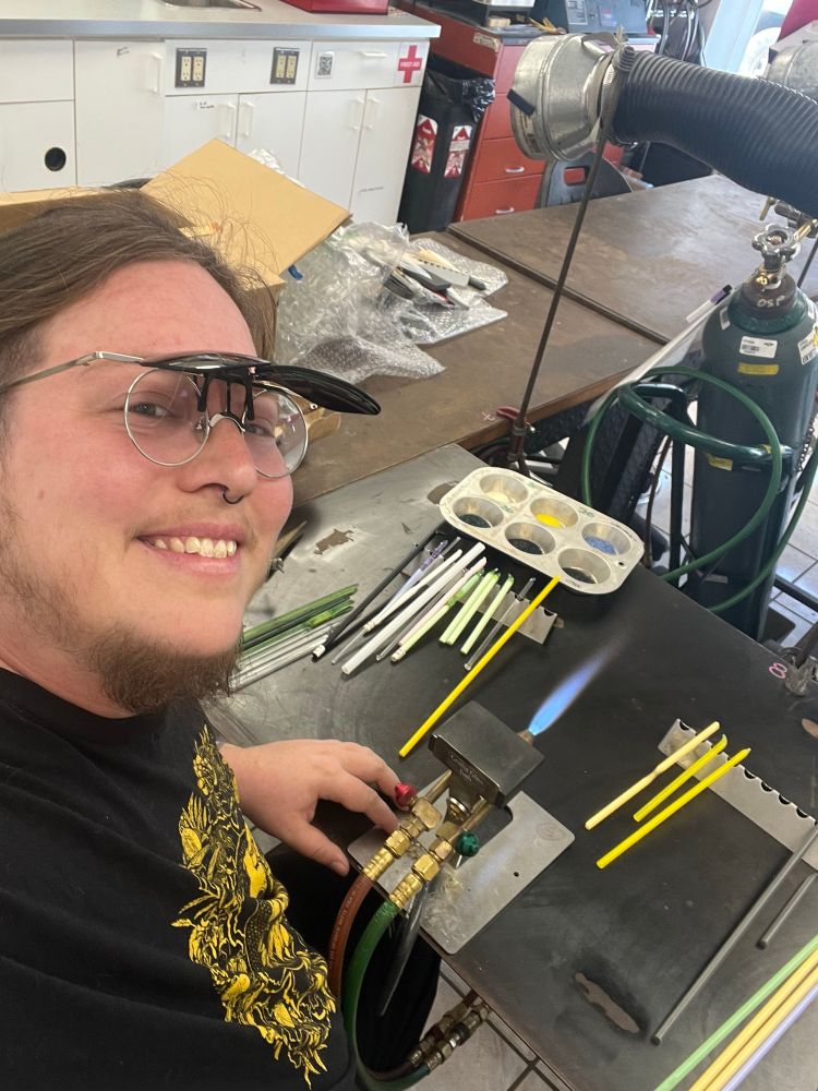 A white person (me) with brown hair, a goatee, glasses (with clip-on shaded lenses flipped up), and a black and yellow shirt smiles as they sit behind a lit torch with rods of glass arrayed on the table in front of them. The flame of the glass torch is blue. 
