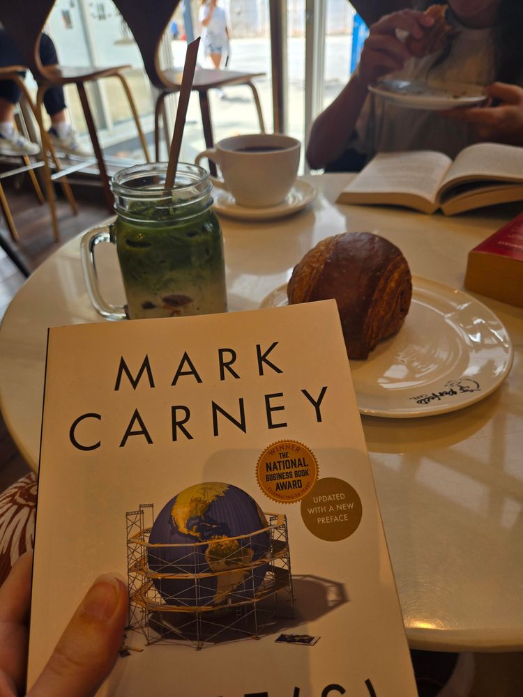 Mark Carney's book in front of a chocolate croissant and an iced matcha latte in a Vancouver café
