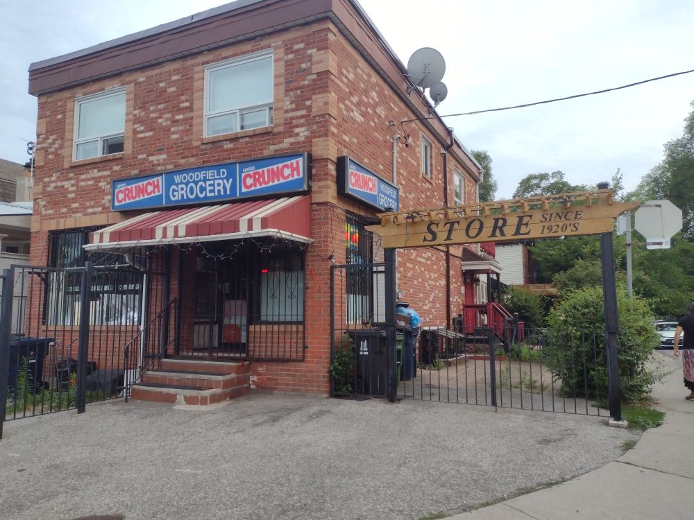 A convenience store located in the first floor of a two-storey early 1900's mixed-use building. It is part of a North American residential neighborhood. The sign the door says "Woodfield Grocery" and is flanked by logos of Nestle Crunch chocolate bars. A secondary wooden sign above a decorative metal gate at the side of the building says "STORE since 1920's".