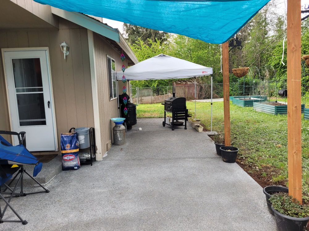 The finished patio with 4x4s installed supporting a teal shadesail in the foreground. Moving along the house there are various plants, a bbq, and a canopy all decorating the patio.