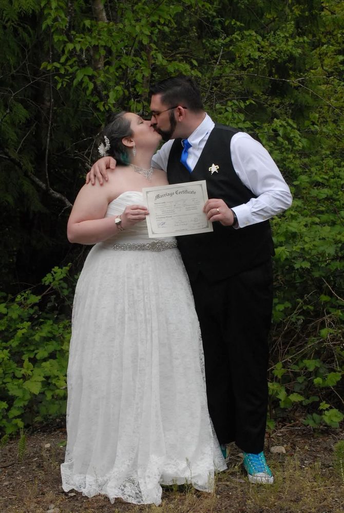 A picture from our wedding as we kiss, holding our marriage certificate. I am to the left in a lacy, white, sleeveless dress. To the right is a man in a black vest and suit pants, sapphire blue tie, and teal high tops. On his shoulder is a Voyager Era comm badge. 