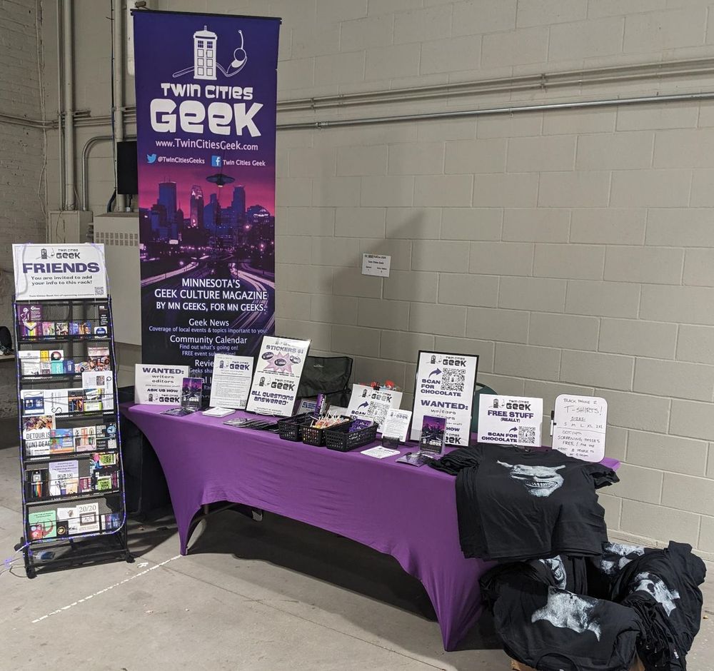 Twin Cities Geek booth at FallCon. It's against the wall. There are lots of signs and an info rack.