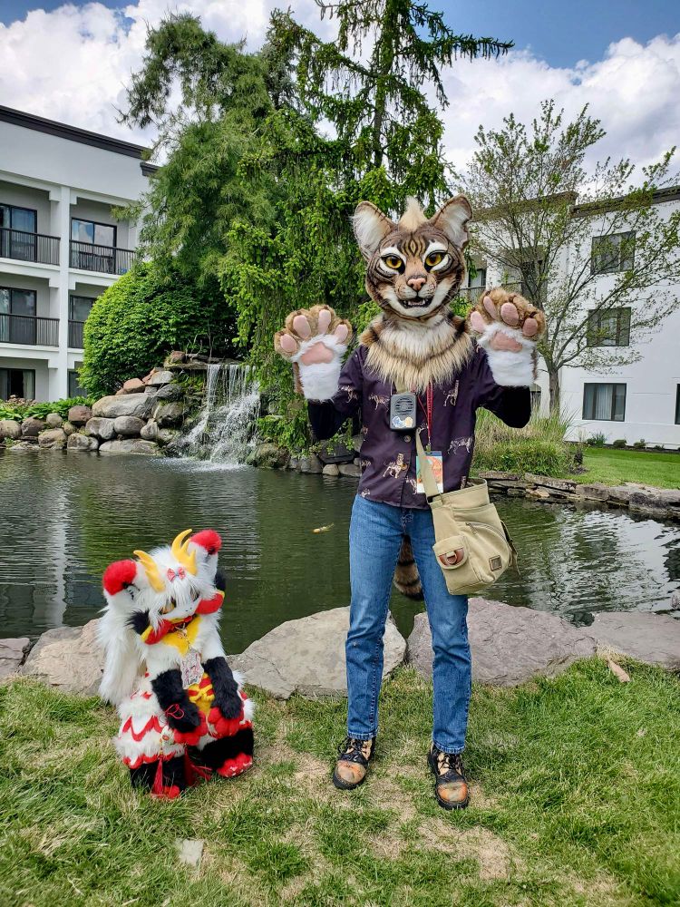 Junebug the tabby cat fursuit and Lumi the noodle dragon plushie posing in front of a koi pond.