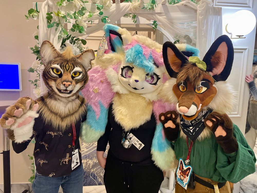 Three fursuits, Junebug the tabby cat, pastel Braixen, and Basil the fox posing in front of a leaf decorated archway. 