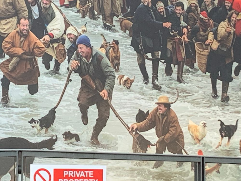 Another part of same mural showing more men, splashing through water with sticks and mutant dogs, including a dog-on-a-stick, all with that strange smile, or is it a grimace?