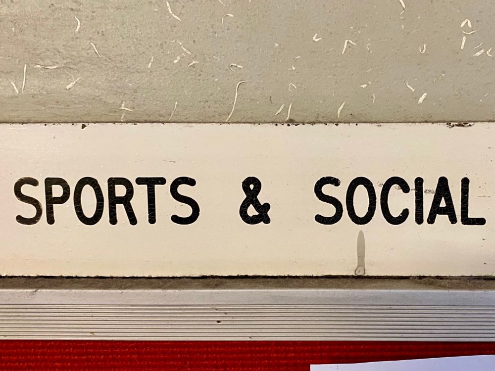 Sign on grey wall. Sports & Social in black all caps on cream background