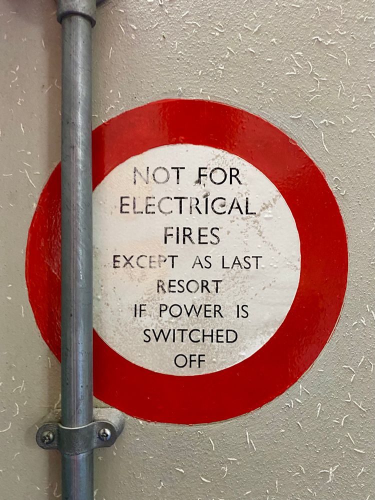 Sign painted on grey wall. Roundel, white centre outlined in red. Inside the white centre is written ‘Not for electrical fires except as last resort if power is switched off’ All caps, sans serif. 1960s?