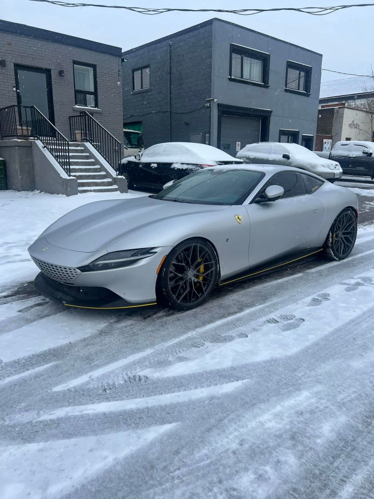 Silver Ferrari Roma parked elegantly in a snowy urban setting, featuring black wheels with yellow accents. A sleek grand tourer contrasting against the winter backdrop