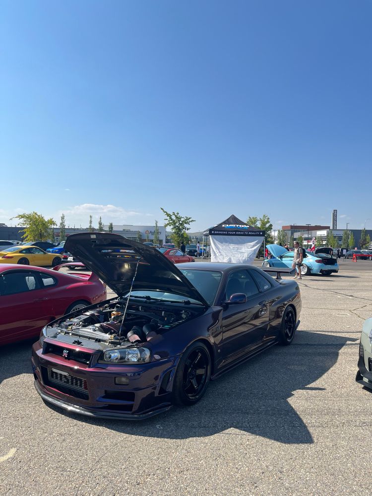 A breathtaking Nissan Skyline R34 GTR finished in the iconic Midnight Purple III stands proudly under the sun at a car meet. Its hood is open, revealing a meticulously detailed engine bay, while its deep, iridescent paint shimmers between shades of purple and black. The car’s aggressive stance is complemented by sleek black wheels, surrounded by a vibrant display of other high-performance vehicles and a clear blue sky above.
