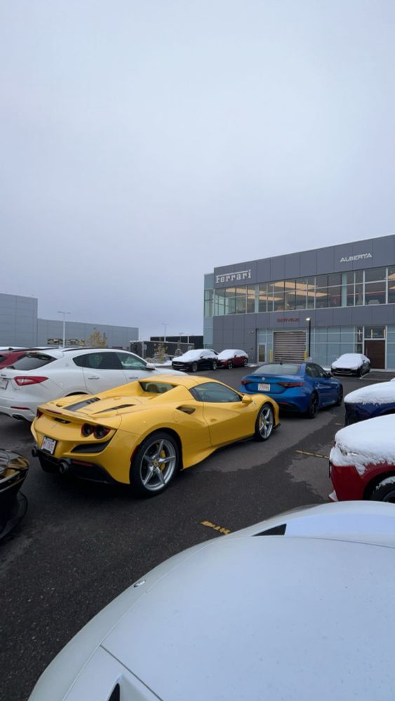 A striking yellow Ferrari F8 Spider sits confidently in a snowy parking lot outside a Ferrari dealership, its vibrant color contrasting sharply with the white snow and muted tones of surrounding cars. A Maserati SUV and a blue sports car add a touch of luxury to the scene, with the sleek Ferrari dealership in the background completing the picture of automotive perfection.