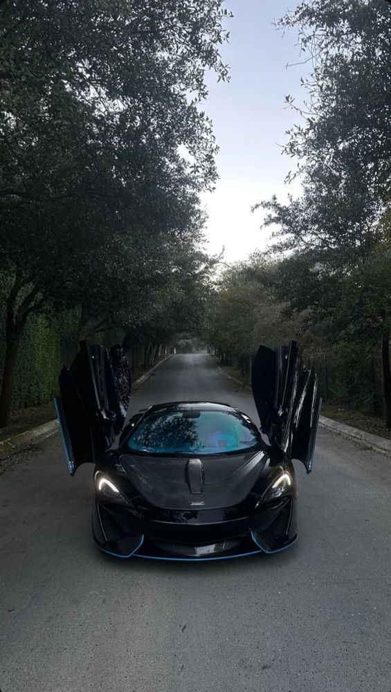 Black McLaren with butterfly doors fully open, parked on a serene, tree-lined road. A subtle blue accent on the front splitter enhances its sleek, futuristic look.