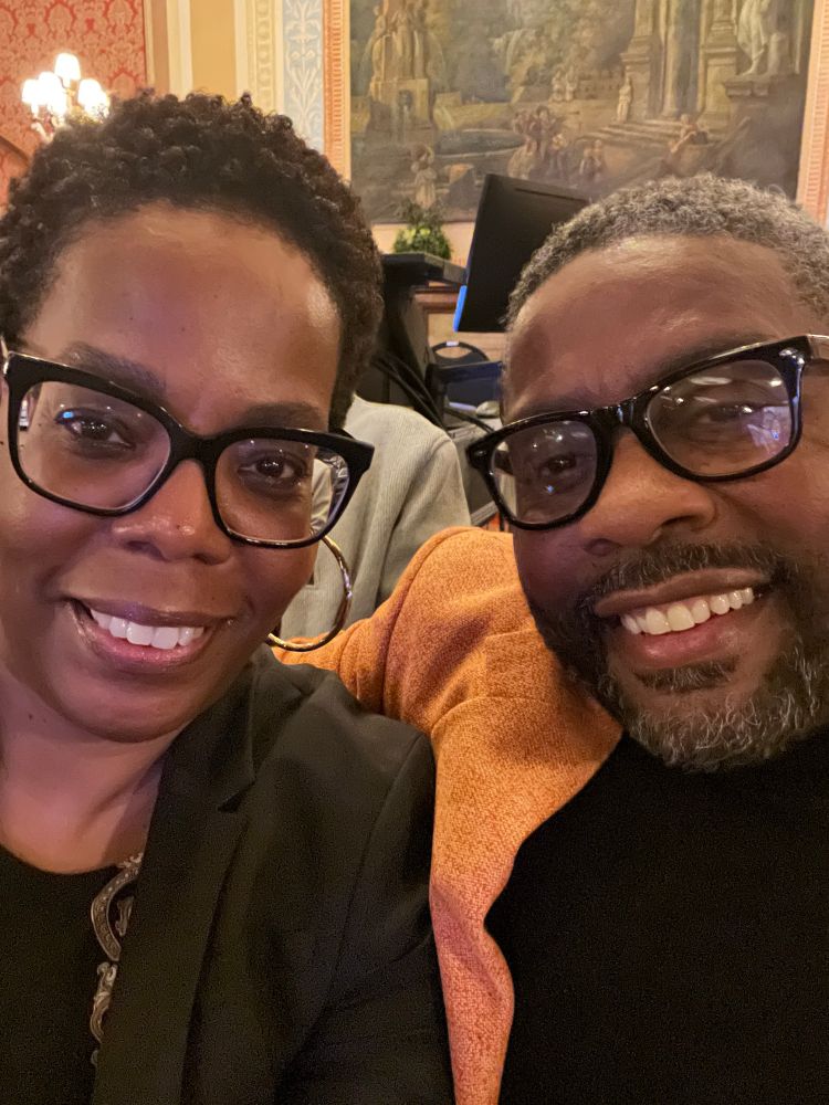 A woman wearing black framed glasses a black jacket and gold hoop earrings. A man wearing black framed glasses, a black sweater and orange blazer. 