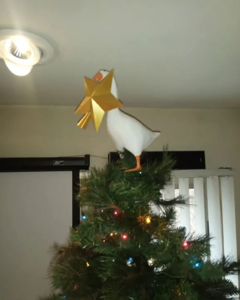 Ganso parado en la cima de un pino con adornos navideños sosteniendo la estrella haciendo alusión al videojuego "Untitled Goose Game".