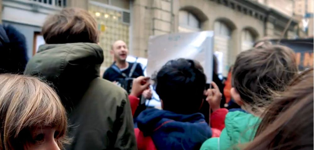 Image avec plusieurs enfants de dos, dont un portant une pancarte, et Reno Bistan qui chante en arrière plan!