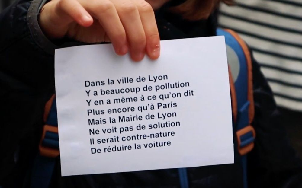 Un enfant tient un papier avec les paroles de la chanson:
Dans le ville de Lyon
Y a beaucoup de pollution
Y en a même à ce qu'on dit
Plus encore qu'à Paris
Mais la mairie de Lyon
Ne voit pas de solution
Il serait contre-nature
De réduire la voiture