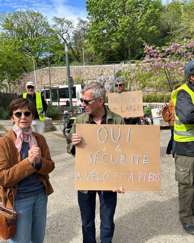 Un homme, à côté d'une femme, porte une pancarte "Oui à la sécurité à vélo et à pieds"
Derrière un autre homme porte une pancarte "Pour des trottoirs plus larges"
