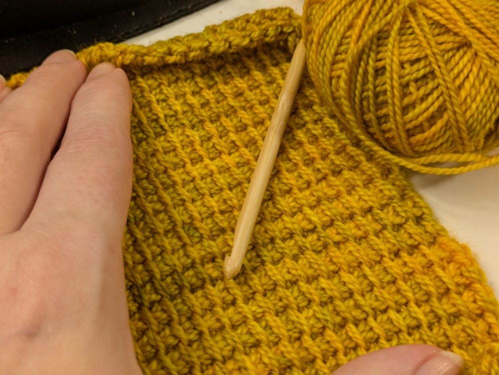 A yellow swatch of Tunisian crochet, made with fingering weight yarn held double. My left hand is spreading the fabric to keep it from rolling too much while I take the photo. 